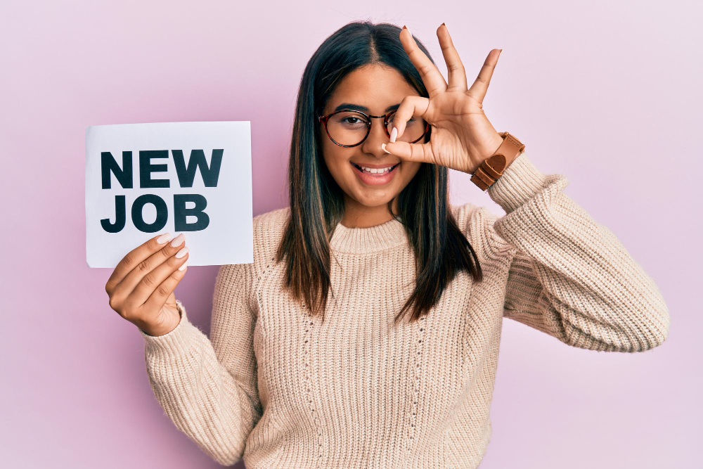 Young latin girl holding new job message on paper smiling happy doing ok sign with hand on eye looking through fingers